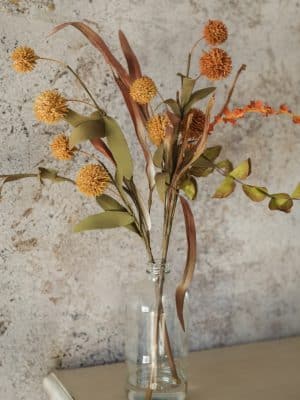 Close-up of mixed fall florals with spiky orange blooms, earthy greens, and rustic tones in a clear vase.