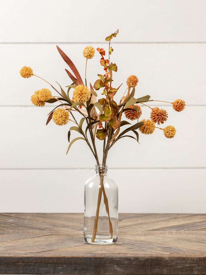 Mixed fall floral arrangement with orange pom pom blooms and autumn leaves styled in a glass bottle vase.