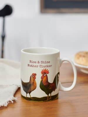 Stoneware mug on a wooden table with hen and rooster design and phrase “Rise & Shine Mother Clucker.”