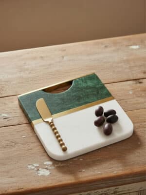 Rounded square emerald wood and white marble serving board with brass inlay, shown on a wood table with a small spreader and olives.