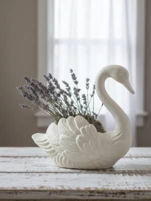 An antique white cast iron swan container is styled on a rustic table holding lavender stems in a softly lit room.