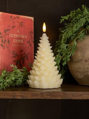 Cream 3D balsam tree flameless candle glowing beside greenery and a red Christmas book