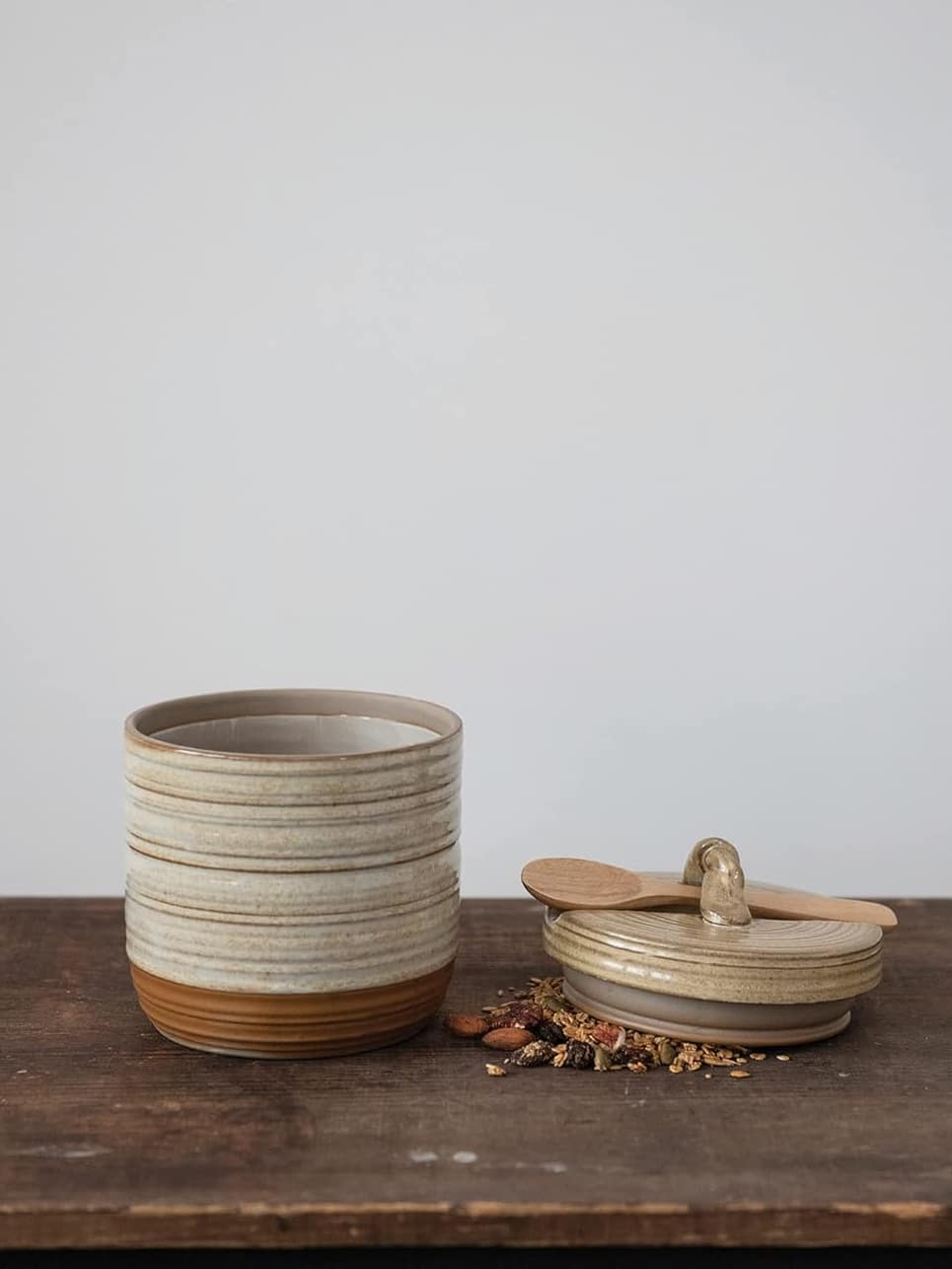 Stoneware jar with tan reactive glaze shown with the lid off beside it and a wooden spoon, styled on a dark surface.