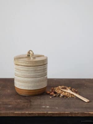 Reactive glaze stoneware lidded jar with a knotted loop handle and matching wooden spoon on a dark tabletop.