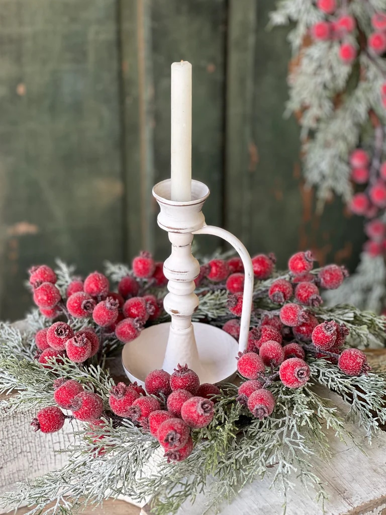 Distressed white metal chamberstick candle holder displayed inside a frosted greenery and red berry wreath.