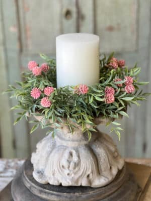 Pink Thimble Buds Candle Ring with soft pink buds and layered greenery styled around a white pillar candle on a carved pedestal.