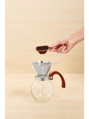 Hand pouring ground coffee into stainless steel filter atop glass pour-over carafe with bamboo handle