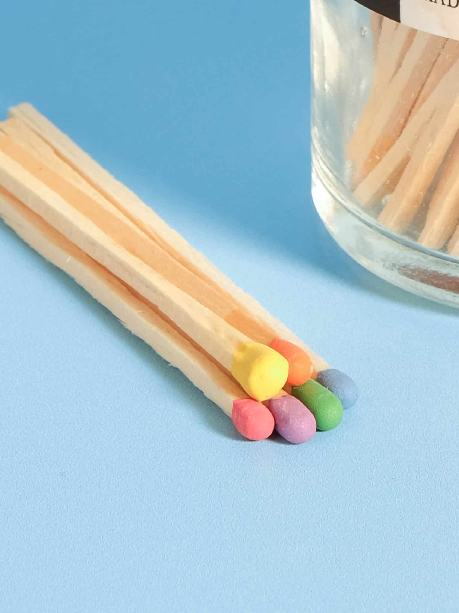 Close-up image of a vibrant array of primary rainbow-colored matchsticks, including red, orange, yellow, green, blue, and purple, neatly arranged against a light blue background.