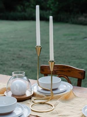 Gold Loop Taper Candle Holder holding two white taper candles, alongside the Wood & Glass Hurricane Candle Holder Small with a white ball candle, all set on a dining table with white plates.