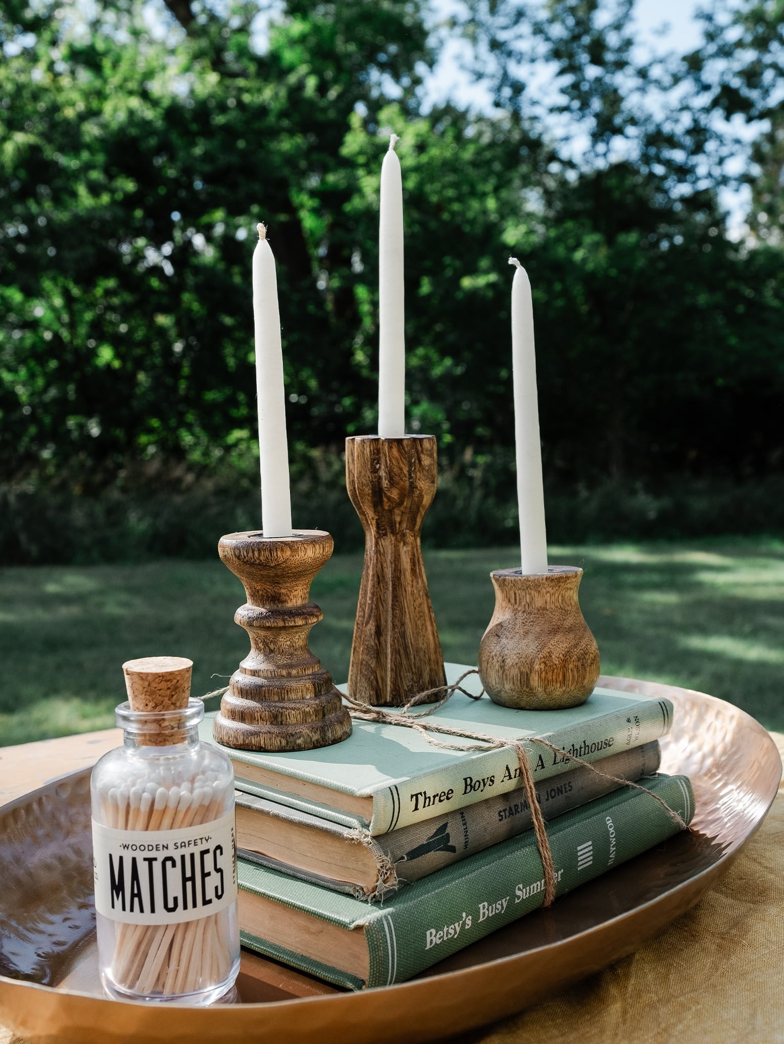 A styled vignette featuring Dual Skinny & Standard Taper Candle Holders, Decorative Vintage Books Set – Cool Colors, Mini Apothecary Vintage White Matches, Skinny Taper Candles – White, and an Oval Brass Platter on a rustic wooden table.