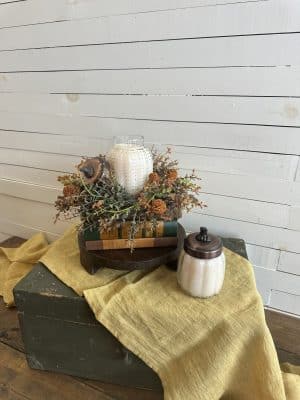 Pumpkin Chata candle and fall floral ring displayed on a wood pedestal with mustard linen table runner