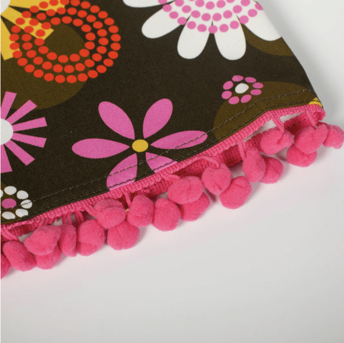 Retro floral tea towel with bold pink, orange, and yellow flowers on a dark brown background and pink pom pom trim.