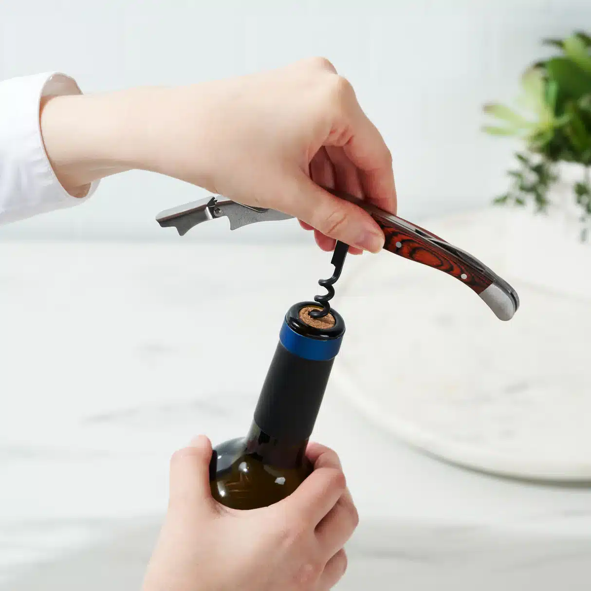 Hand using a wood-handled stainless steel corkscrew to open a wine bottle in a kitchen setting.