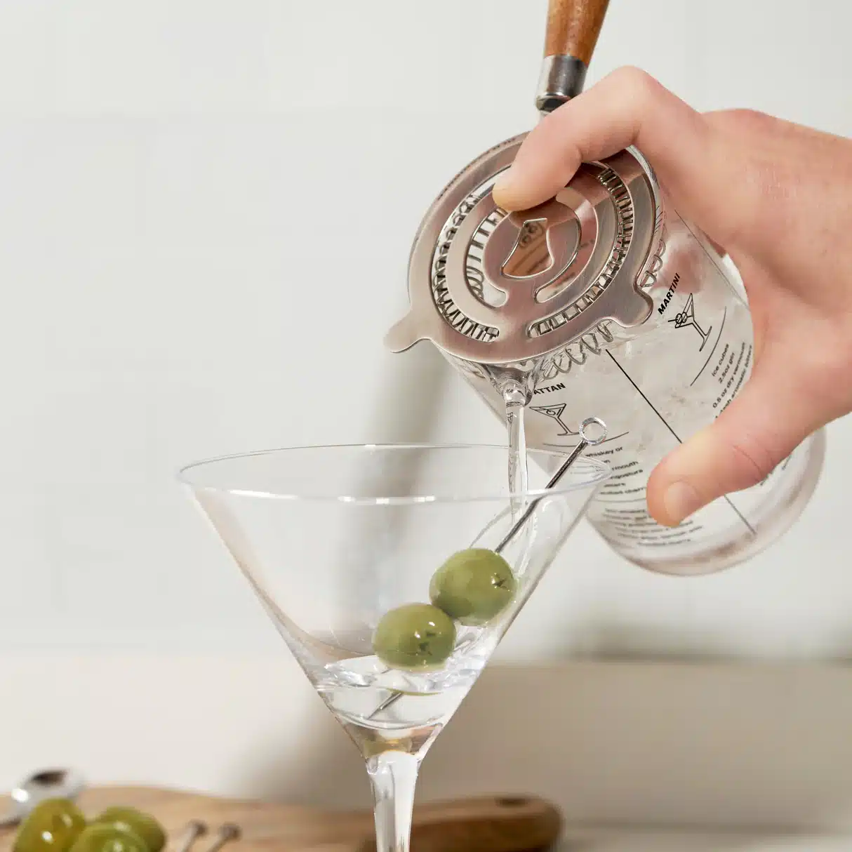 Hand using cocktail strainer to pour a martini into a glass while blocking ice and olives.