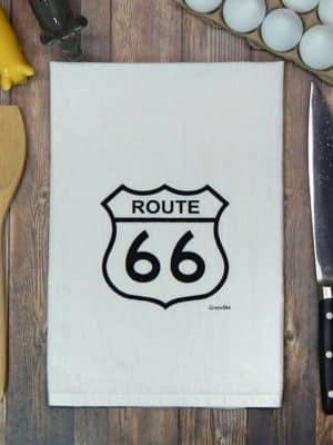 Cotton flour sack tea towel printed with the black Route 66 highway sign, styled on a wooden countertop with kitchen tools.
