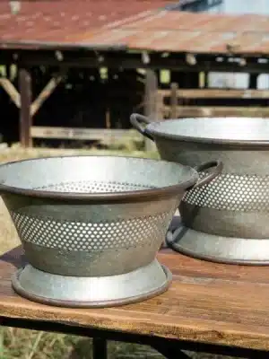 Close-up of two distressed galvanized colander planters with side handles and footed bases, showcasing their rustic finish and repurposed style.
