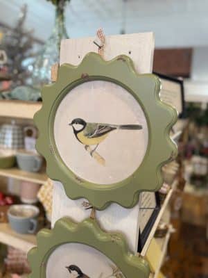 Vintage-style Yellow-Breasted Finch wall art in a distressed scalloped green frame, displayed on a white wood board in a cozy shop setting.