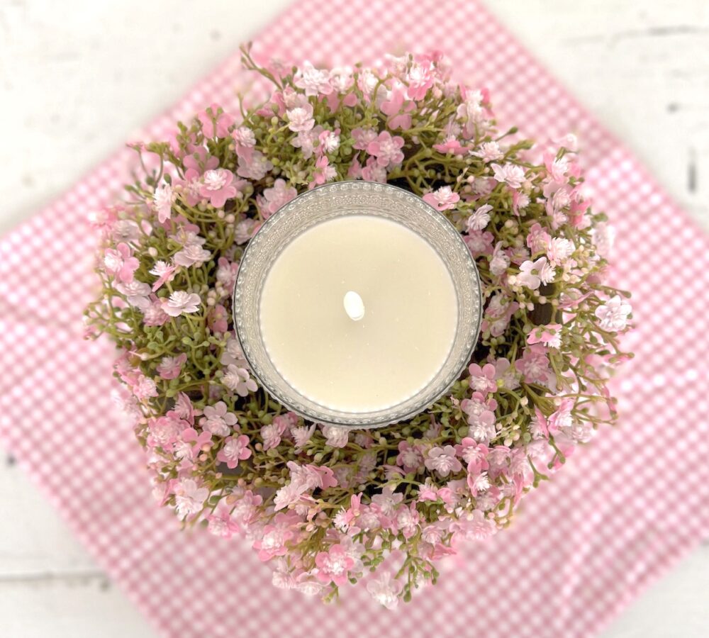 Top view of a clear dotted glass flameless candle nestled inside a Pink Stardust Blooms Candle Ring on a white pedestal bowl over a pink gingham runner.