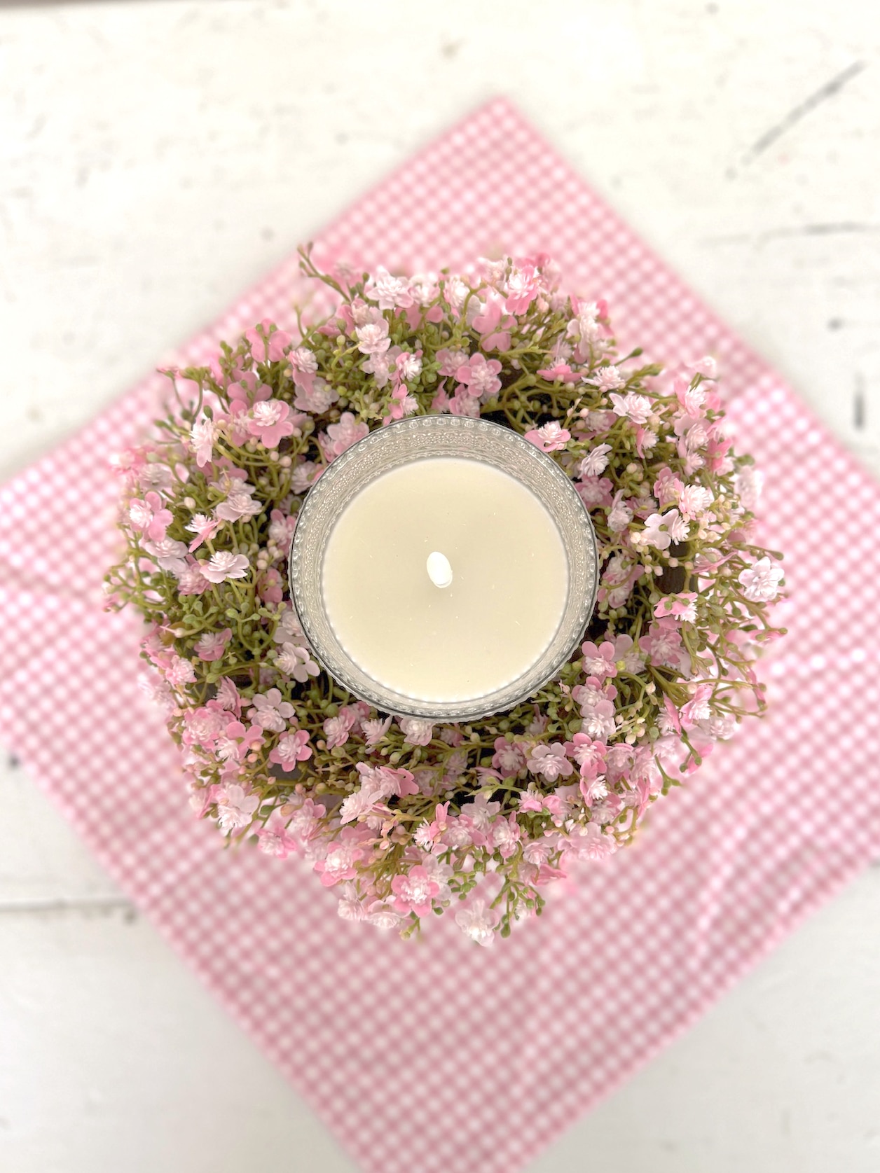 Top view of a clear dotted glass flameless candle nestled inside a Pink Stardust Blooms Candle Ring on a white pedestal bowl over a pink gingham runner.