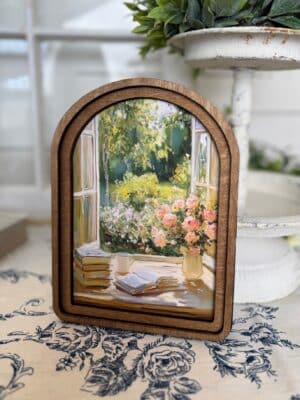 Arched wood framed canvas wall art of an open cottage window with blooming florals, stacked books, and a coffee cup.