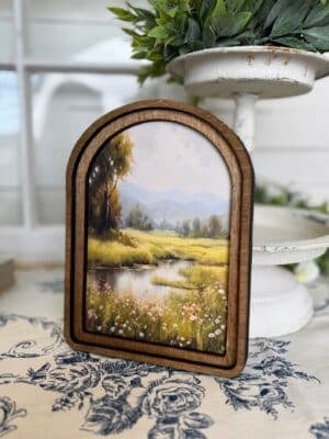Arched wood framed canvas wall art featuring a peaceful stream, wildflowers, and distant hills in soft countryside tones.