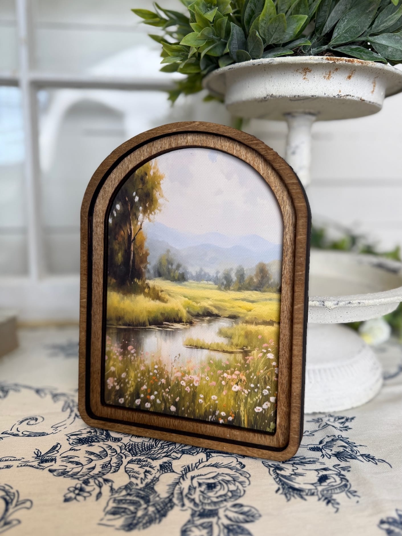 be-made-hays-ks-spring-stream-arched-wall-art Arched wood framed canvas wall art featuring a peaceful stream, wildflowers, and distant hills in soft countryside tones.
