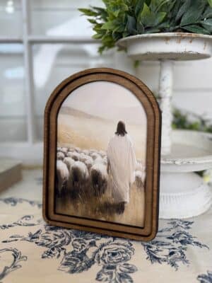 Arched wood framed canvas wall art of Jesus walking among sheep in soft neutral countryside tones.