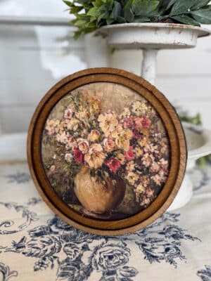 Round wood framed canvas wall art featuring a vintage floral bouquet in a rustic vase with warm aged tones.