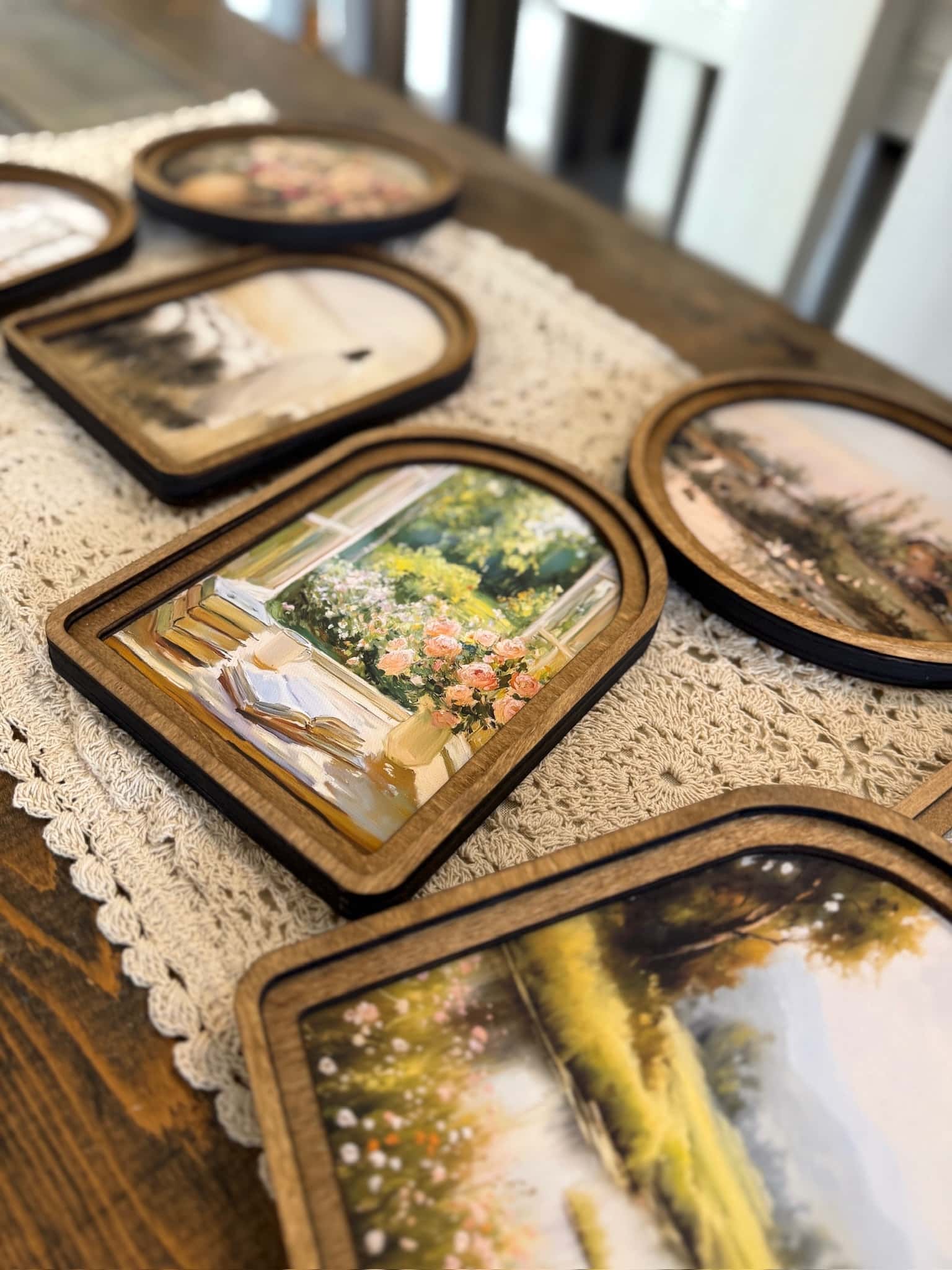 Cottage-style framed canvas wall art featuring florals, countryside scenes, and arched wood frames arranged on wooden table.